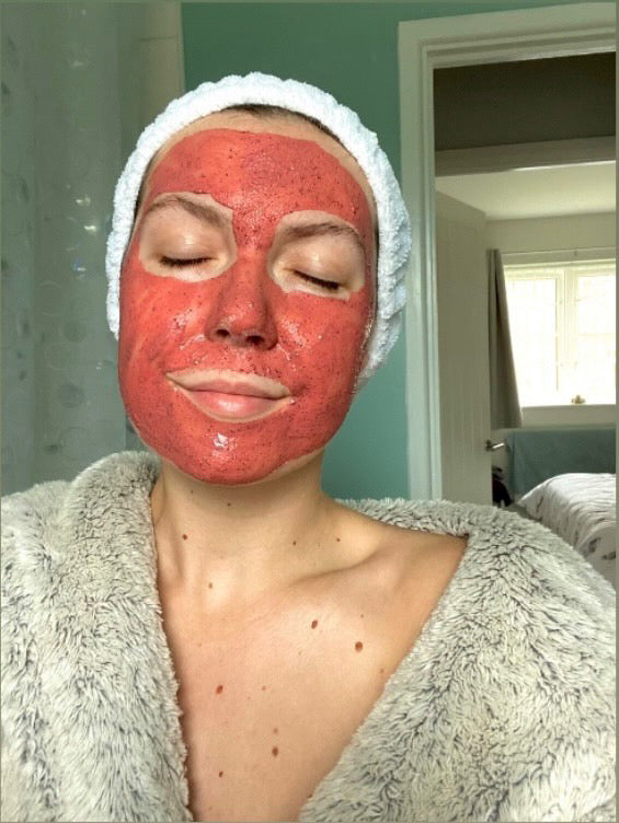 Person with a red facial mask and towel on head in a bathroom setting