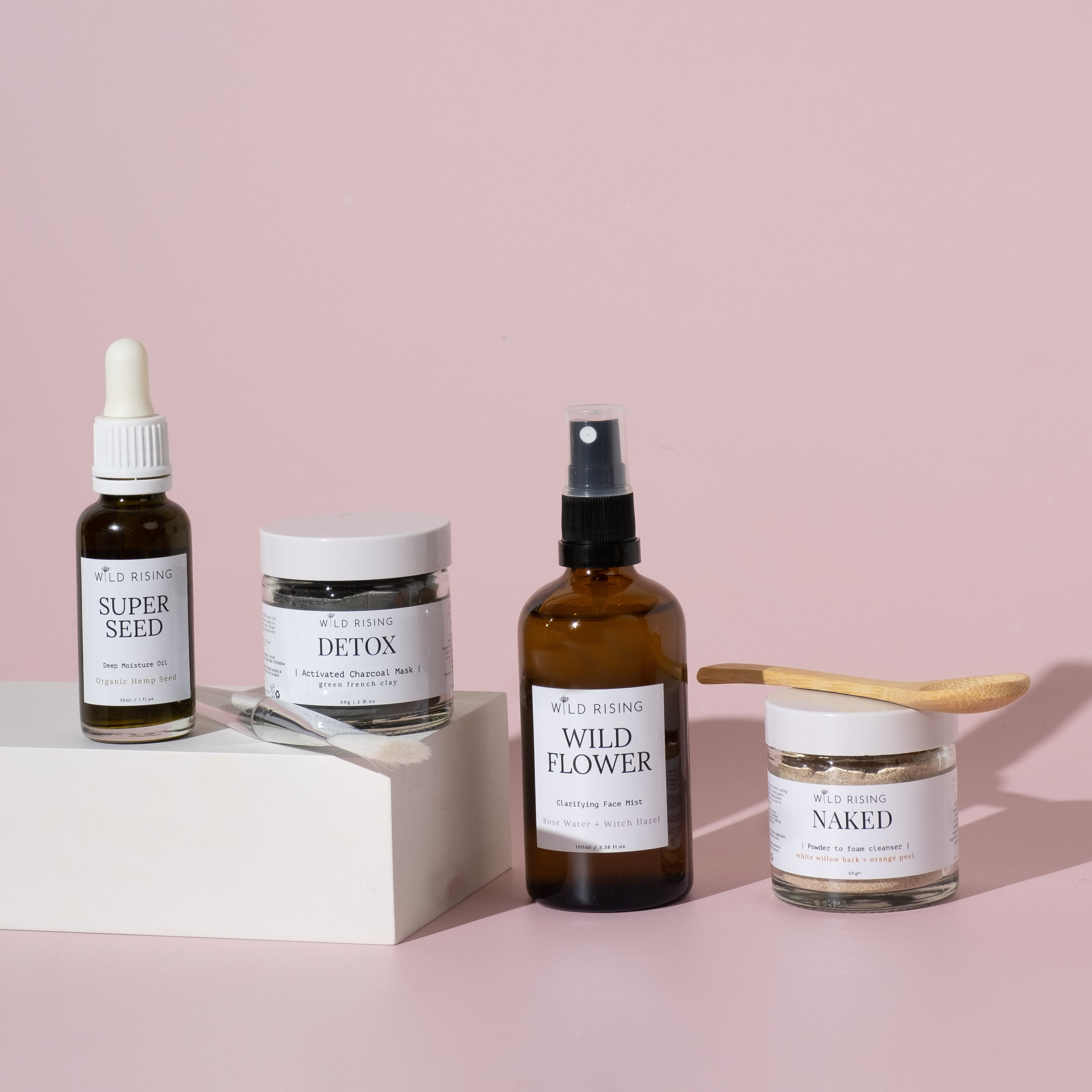 A collection of skincare products including a cleanser, toner, face oil, and face mask, with bottles and containers displayed on a white surface against a pink background.