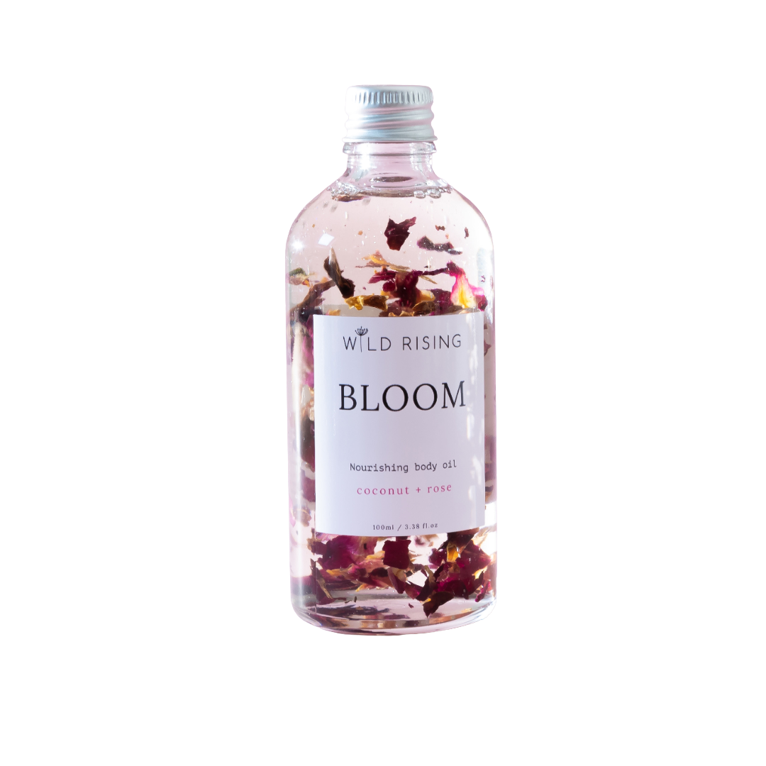 A clear bottle of Wild Rising Bloom nourishing body oil with dried rose petals suspended inside, set against a plain white background.