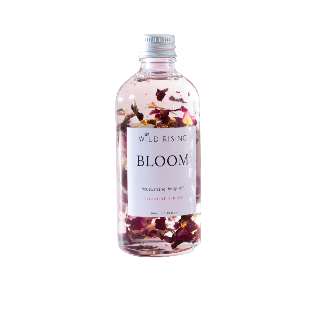 A clear bottle of Wild Rising Bloom nourishing body oil with dried rose petals suspended inside, set against a plain white background.
