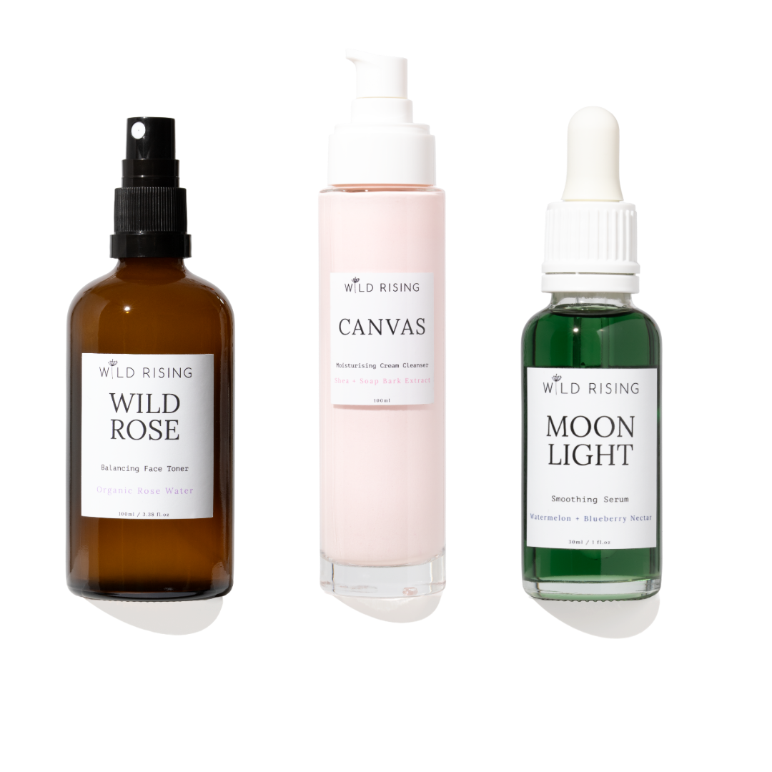 Three skincare products from WILD RISING on a white background: an amber spray bottle labeled Wild Rose, a pale pink pump bottle labeled Canvas, and a green dropper bottle labeled Moon Light.