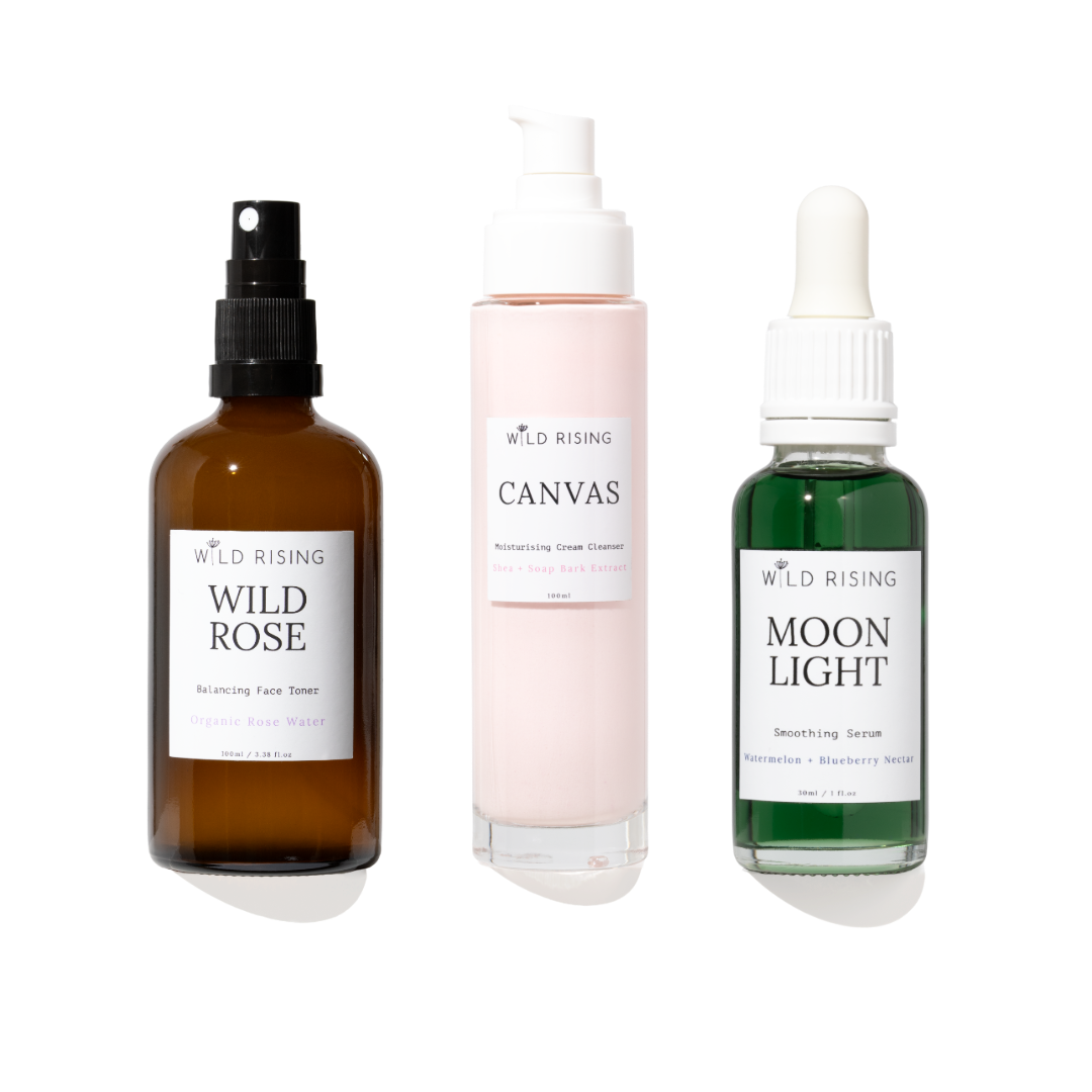 Three skincare products from WILD RISING on a white background: an amber spray bottle labeled Wild Rose, a pale pink pump bottle labeled Canvas, and a green dropper bottle labeled Moon Light.