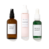 Three skincare products from WILD RISING on a white background: an amber spray bottle labeled Wild Rose, a pale pink pump bottle labeled Canvas, and a green dropper bottle labeled Moon Light.