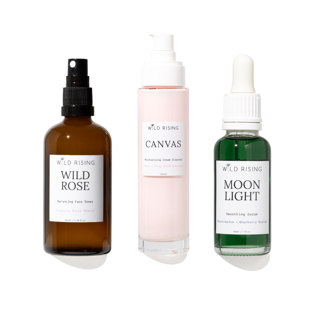 Three skincare products from Wild Rising on a white background: a brown bottle of Wild Rose toner, a pale pink pump bottle of Canvas moisturizing cream, and a green dropper bottle of Moon Light smoothing serum.