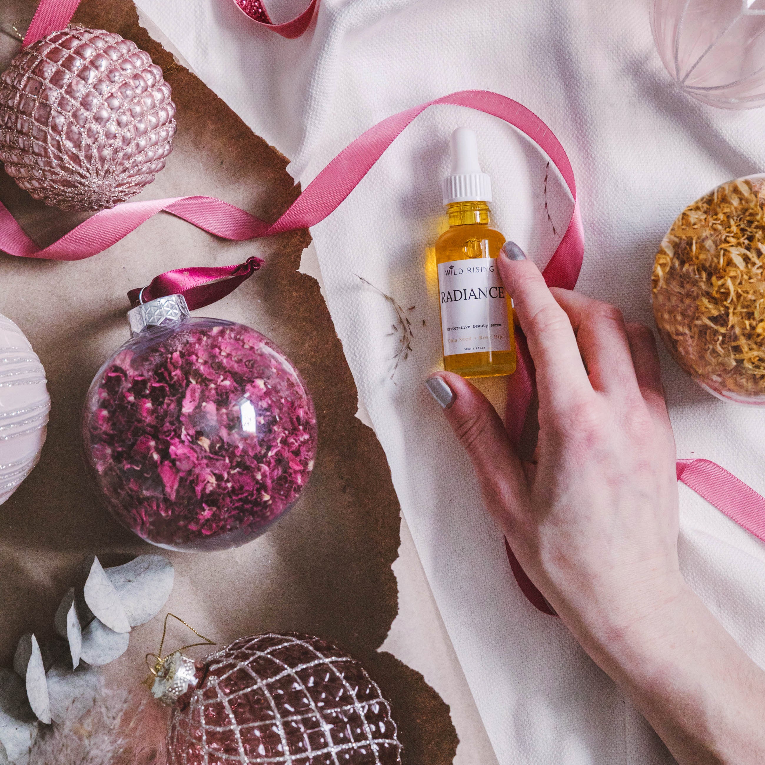 Hand holding a bottle of perfume next to decorative Christmas ornaments on a white surface with pink ribbons.