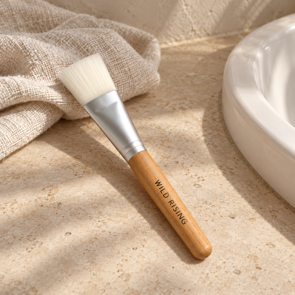 A makeup brush with a light wood handle and white bristles lies on a beige stone surface next to a white ceramic bowl and a textured beige cloth. The handle is engraved with the brand name 'WILD RISING'.