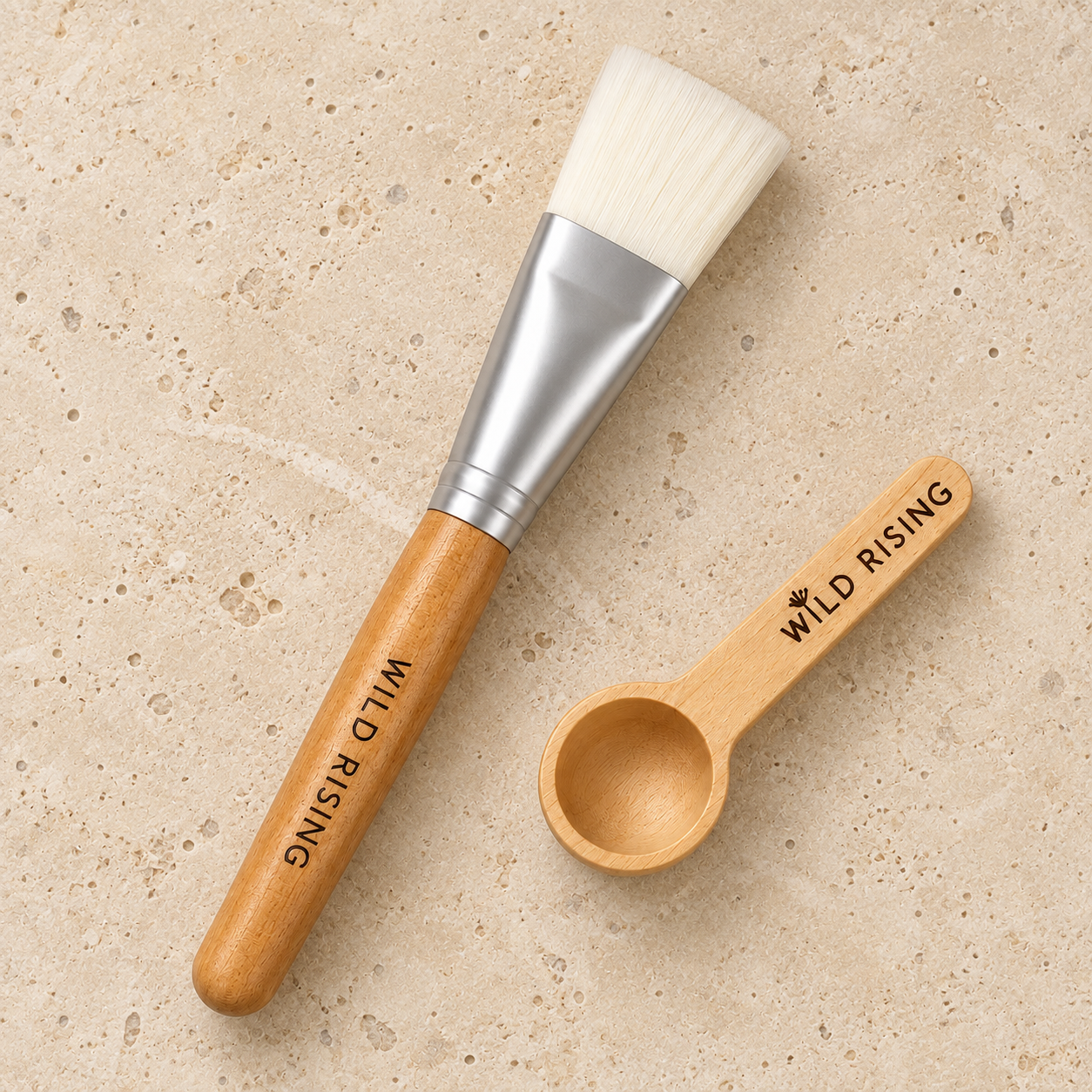 A wooden-handled makeup brush with white bristles and a small matching wooden scoop, both engraved with the brand name 'WILD RISING,' placed on a light beige stone surface.