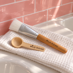 A wooden-handled makeup brush and a small wooden scoop with the brand name 'WILD RISING' engraved on both, resting on a white textured towel in front of pink subway tile.
