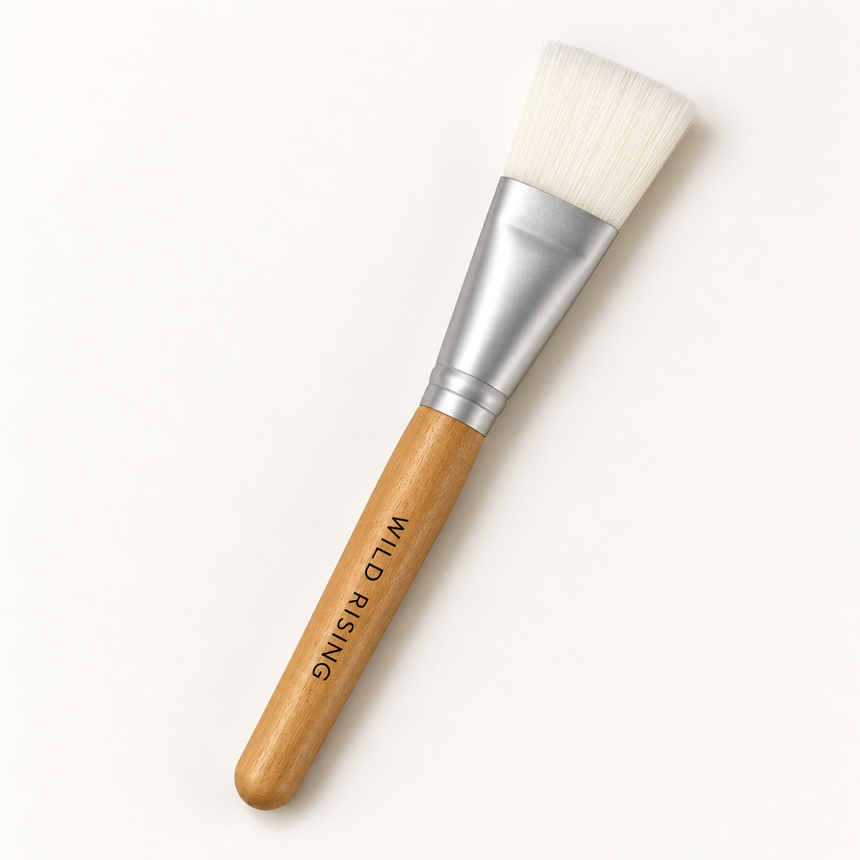 A flat makeup brush with white bristles, a silver metal ferrule, and a light wood handle. The handle is printed with the brand name 'WILD RISING'.