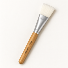 A flat makeup brush with white bristles, a silver metal ferrule, and a light wood handle. The handle is printed with the brand name 'WILD RISING'.