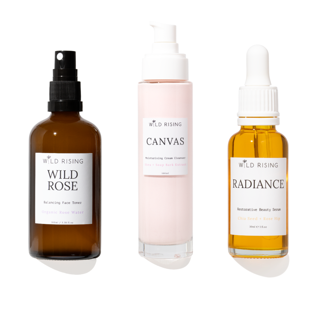 Three skincare products from Wild Rising on a white background: Wild Rose Balancing Face Toner in an amber spray bottle, Canvas Moisturising Cream Cleanser in a pale pink pump bottle, and Radiance Serum in an amber dropper bottle.