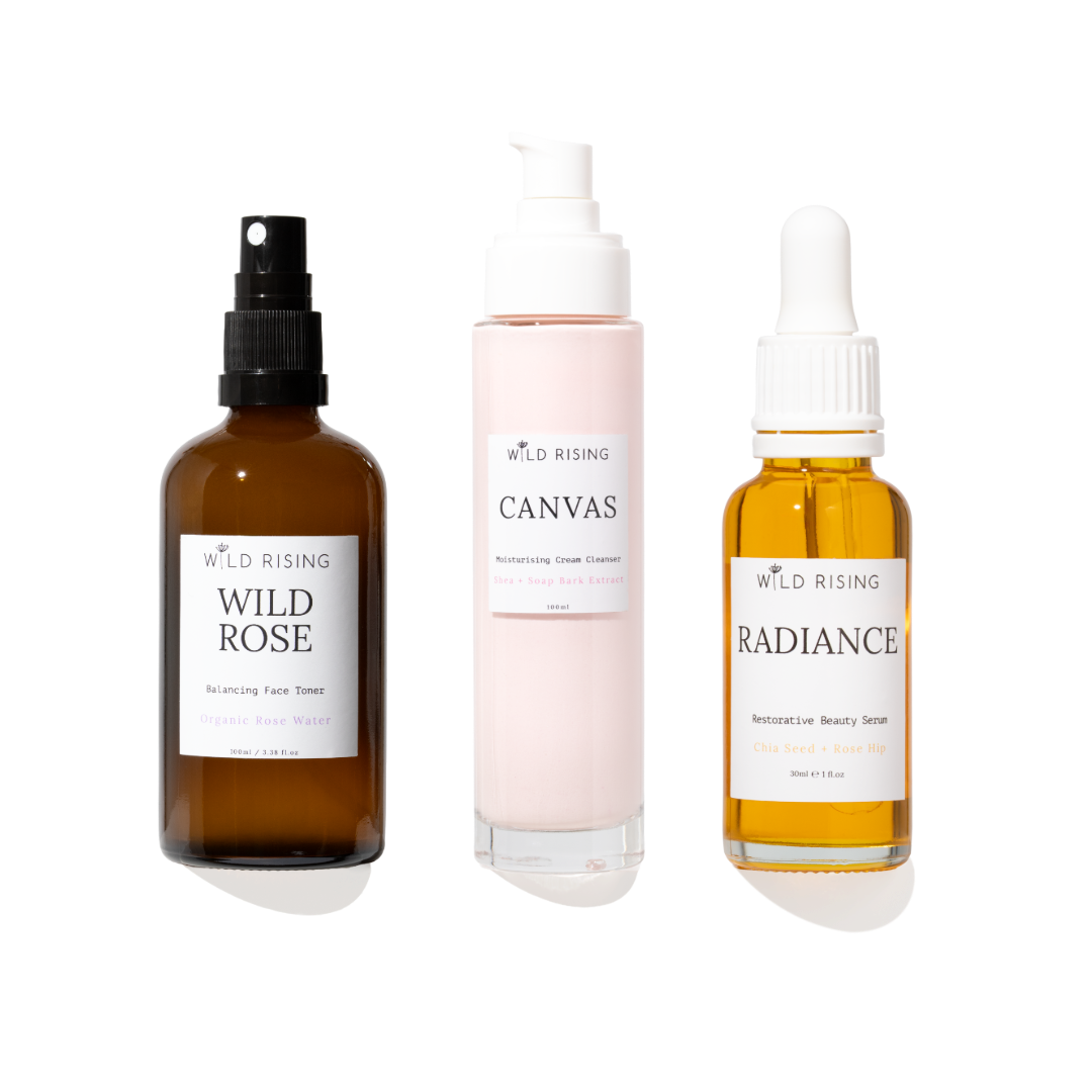 Three skincare products from Wild Rising on a white background: Wild Rose Balancing Face Toner in an amber spray bottle, Canvas Moisturising Cream Cleanser in a pale pink pump bottle, and Radiance Serum in an amber dropper bottle.