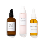 Three skincare products from Wild Rising on a white background: Wild Rose Balancing Face Toner in an amber spray bottle, Canvas Moisturising Cream Cleanser in a pale pink pump bottle, and Radiance Serum in an amber dropper bottle.