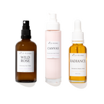 Three Wild Rising skincare bottles on a white background: an amber spray bottle labeled Wild Rose, a pale pink pump bottle labeled Canvas, and an amber dropper bottle labeled Radiance.
