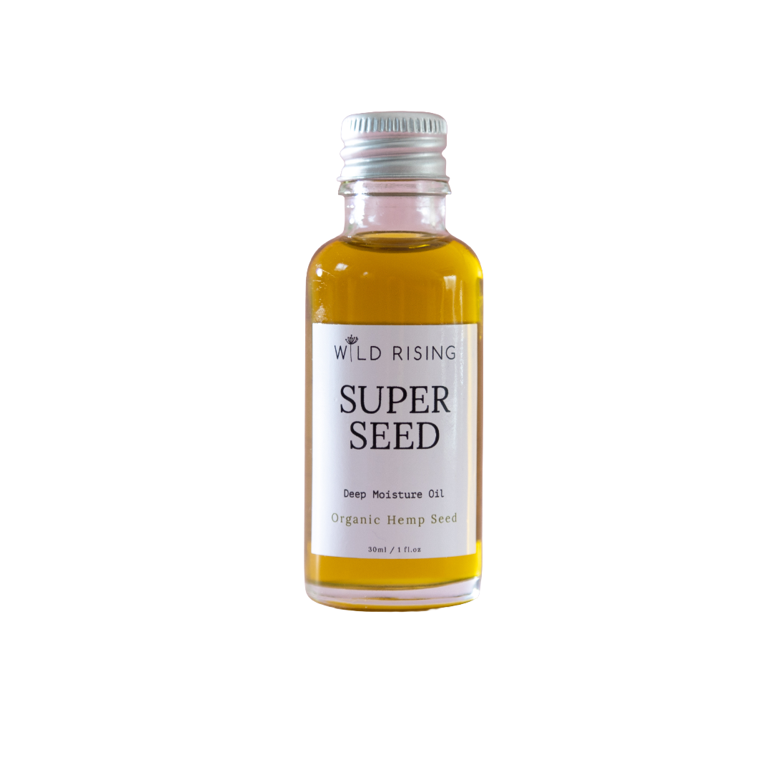 A small glass bottle of Wild Rising Super Seed Deep Moisture Oil filled with golden-yellow oil, featuring a white label and a silver metal cap, set against a plain white background.