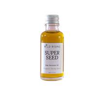 A small glass bottle of Wild Rising Super Seed Deep Moisture Oil filled with golden-yellow oil, featuring a white label and a silver metal cap, set against a plain white background.