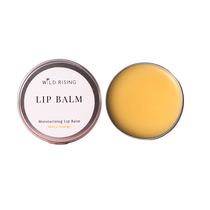 Two round metal tins of Wild Rising Lip Balm on a plain white background, one closed with a pale pink label reading 'LIP BALM Moisturising Lip Balm Zesty Orange' and one open showing the balm inside, which is a warm yellow-orange color.