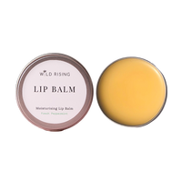 Two round metal tins of Wild Rising Lip Balm on a white background, one closed with a pale pink label and one open showing the yellow balm inside.