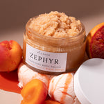An open glass jar of Zephyr Energing Body Polish by Wild Rising, filled with a light tan scrub, styled with apricots, a blood orange slice, and ice cream on a warm beige background.