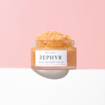 A clear glass jar of Wild Rising Zephyr Energising Body Polish with a white label, filled with a peachy-orange scrub and topped with coarse scrub, set against a split pink and white background.