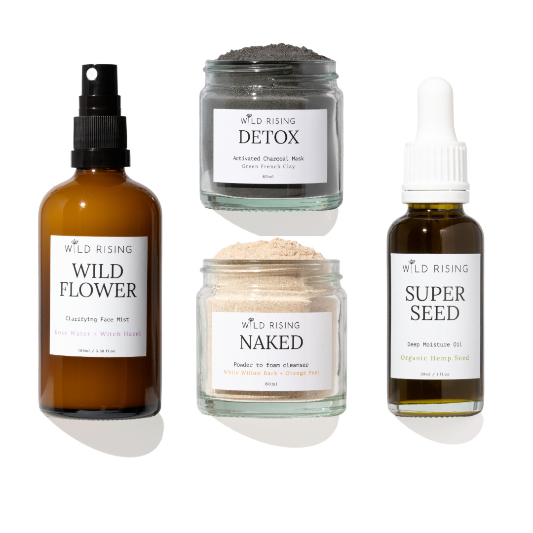 Four skincare products from Wild Rising arranged on a white background: a brown spray bottle labeled Wild Flower, a gray detox mask jar labeled Detox, a beige powder cleanser jar labeled Naked, and a dark seed oil bottle labeled Super Seed.