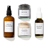 Four skincare products from Wild Rising arranged on a white background: a brown spray bottle labeled Wild Flower, a gray detox mask jar labeled Detox, a beige powder cleanser jar labeled Naked, and a dark seed oil bottle labeled Super Seed.