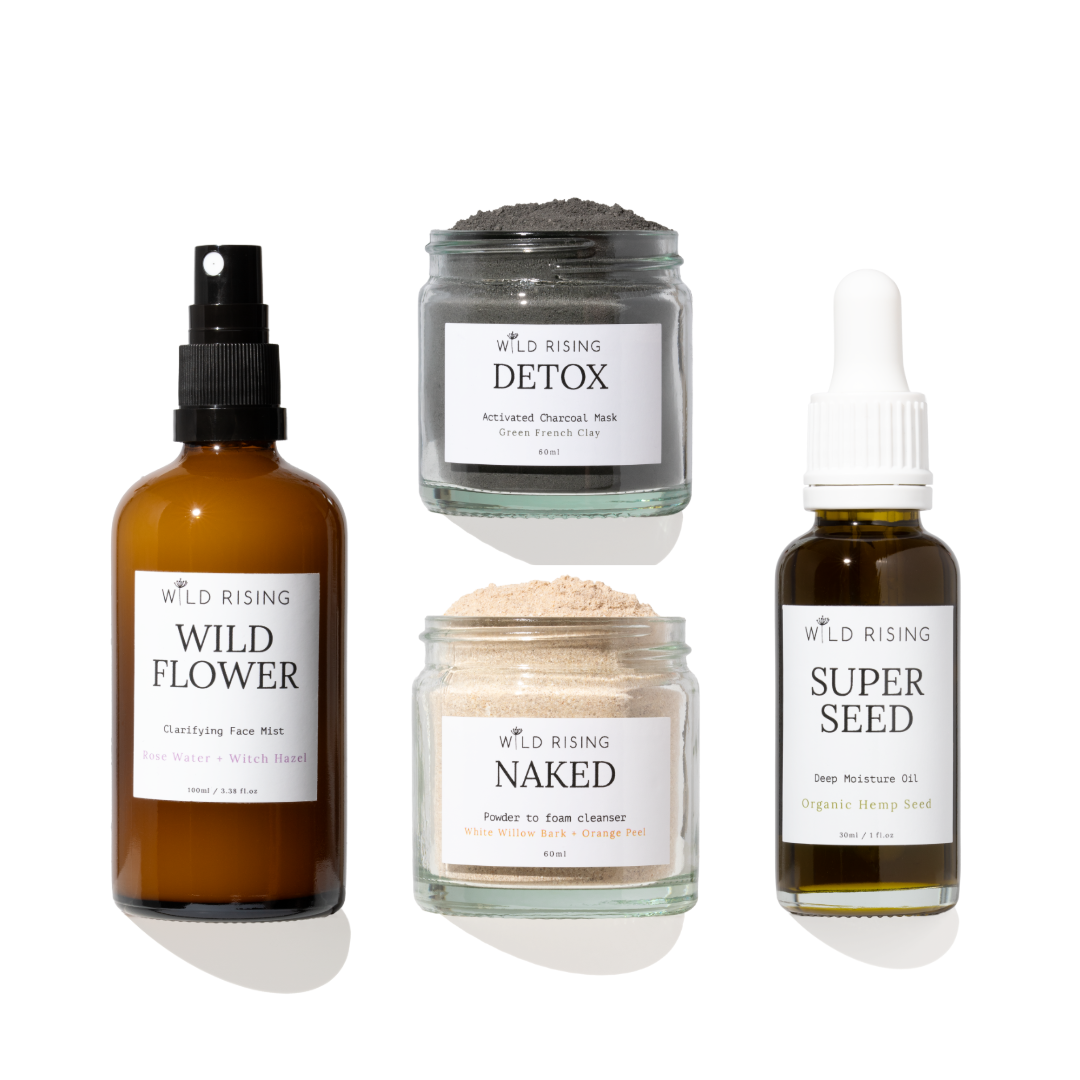 Four skincare products from Wild Rising arranged on a white background: a brown spray bottle labeled Wild Flower, a gray detox mask jar labeled Detox, a beige powder cleanser jar labeled Naked, and a dark seed oil bottle labeled Super Seed.