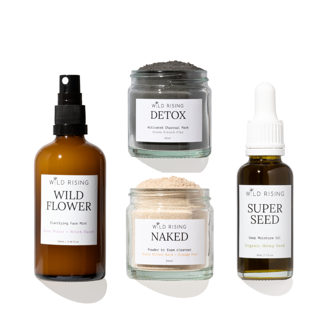 Four skincare products from Wild Rising arranged on a white background: a brown spray bottle labeled Wild Flower, a gray detox mask jar labeled Detox, a beige powder cleanser jar labeled Naked, and a dark seed oil bottle labeled Super Seed.