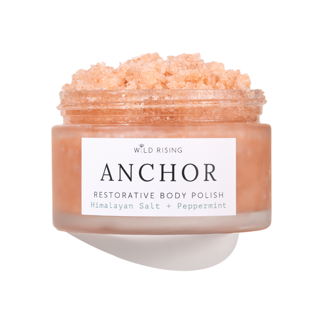 A jar of Wild Rising Anchor Restorative Body Polish, filled with a peach-colored crystalline scrub, shown against a clean white background with a soft shadow.