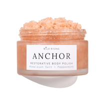 A jar of Wild Rising Anchor Restorative Body Polish, filled with a peach-colored crystalline scrub, shown against a clean white background with a soft shadow.