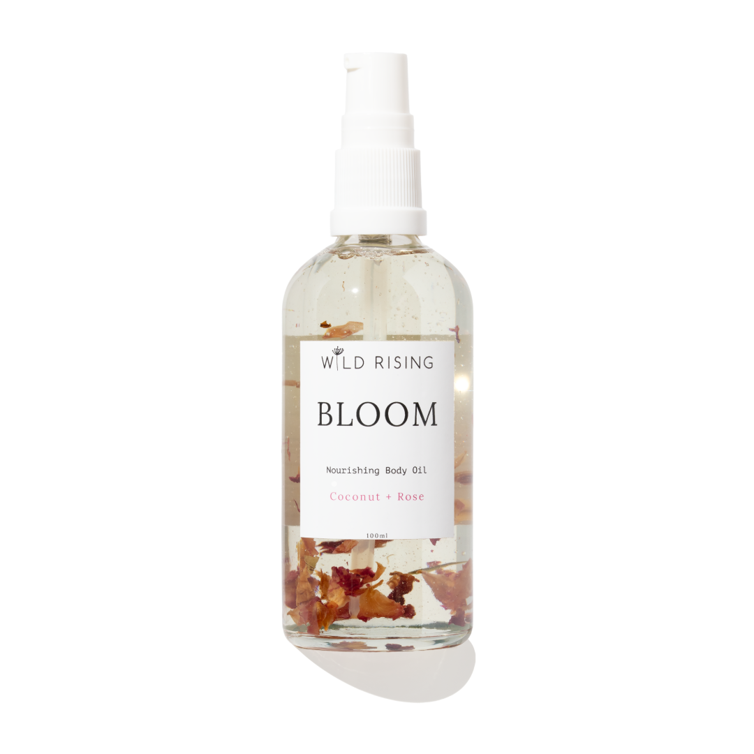 A clear spray bottle of Wild Rising Bloom Nourishing Body Oil with a white pump top, featuring dried coconut and rose botanicals suspended in the clear oil, set against a white background with a soft pink gradient at the bottom.