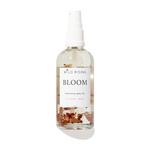 A clear spray bottle of Wild Rising Bloom Nourishing Body Oil with a white pump top, featuring dried coconut and rose botanicals suspended in the clear oil, set against a white background with a soft pink gradient at the bottom.