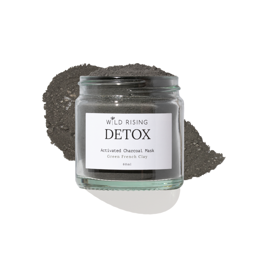 A glass jar of Wild Rising Detox Activated Charcoal Mask, filled with dark green French clay powder and topped with a small pile of the same charcoal mask, set against a clean white background.