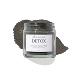 A glass jar of Wild Rising Detox Activated Charcoal Mask, filled with dark green French clay powder and topped with a small pile of the same charcoal mask, set against a clean white background.