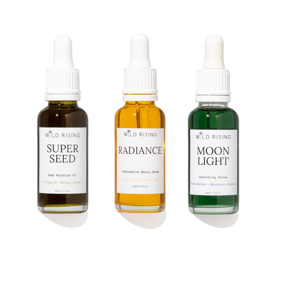 Three Wild Rising dropper bottles arranged side by side on a white background: Super Seed deep moisture oil in dark amber, Radiance restorative beauty serum in golden amber, and Moon Light smoothing serum in green.