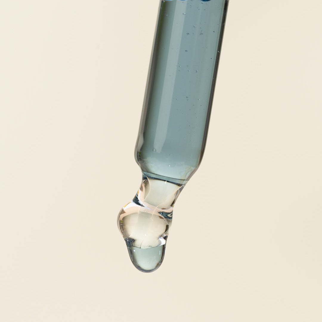 A close-up studio shot of a glass dropper containing a pale blue liquid, with a single clear droplet hanging beneath the bulb against a warm off-white background.