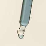 A close-up studio shot of a glass dropper containing a pale blue liquid, with a single clear droplet hanging beneath the bulb against a warm off-white background.