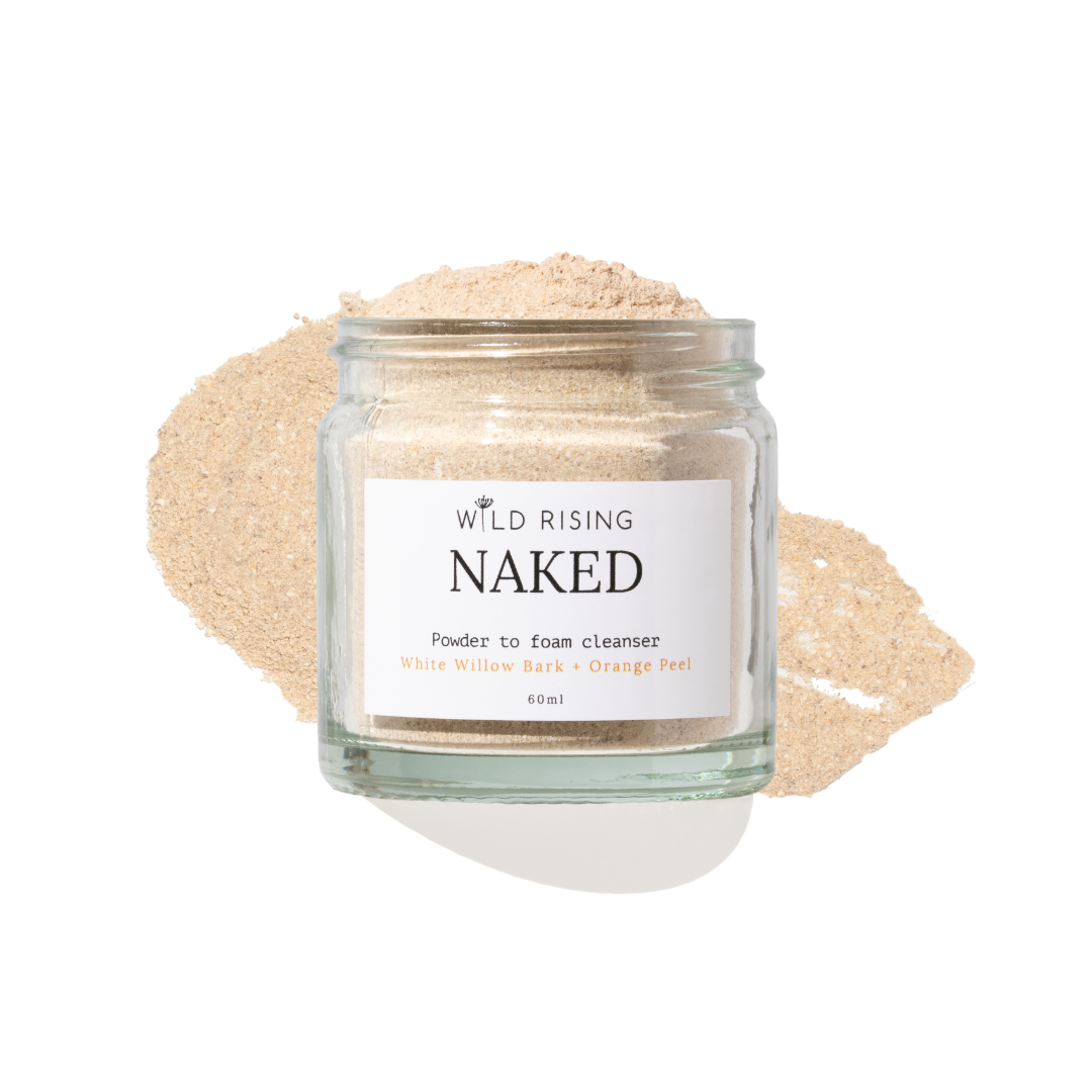 A clear glass jar labeled 'WILD RISING NAKED' containing a light beige powder, set against a matching beige powder smear on a white background.