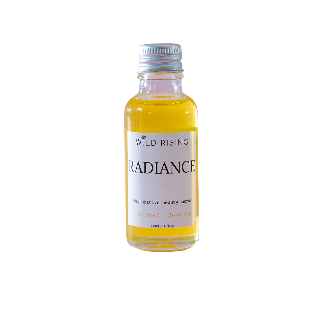 A small glass bottle of Wild Rising Radiance restorative beauty serum with a silver cap, filled with yellow liquid, on a white background.