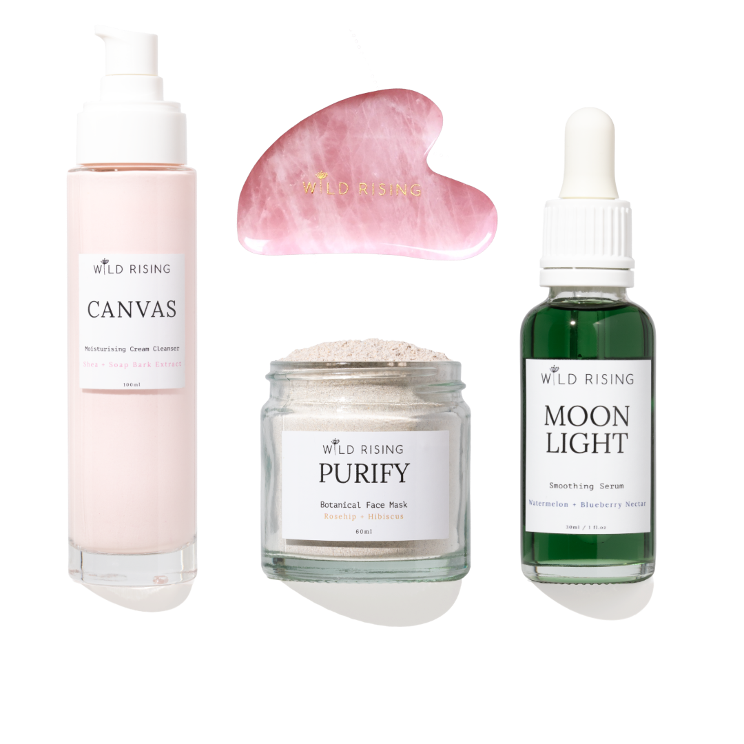 Four skincare products from Wild Rising arranged on a white background: a pale pink pump bottle labeled Canvas, a pink gua sha massage tool, a clear jar labeled Purify Botanical Face Mask, and a dark green dropper bottle labeled Moon Light Smoothing Serum