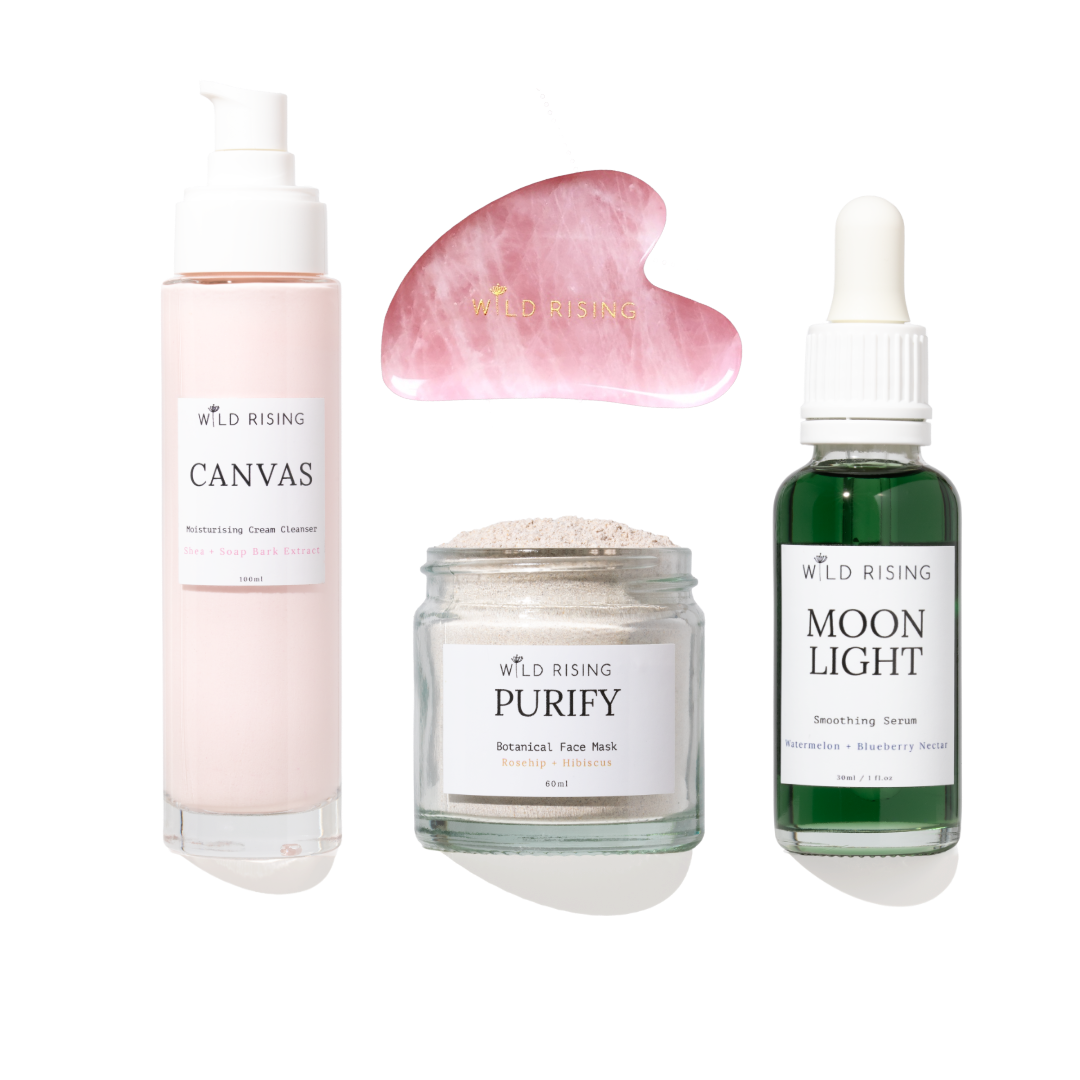 Four skincare products from Wild Rising arranged on a white background: a pale pink pump bottle labeled Canvas, a pink gua sha massage tool, a clear jar labeled Purify Botanical Face Mask, and a dark green dropper bottle labeled Moon Light Smoothing Serum