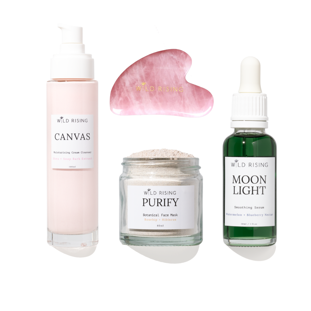 Four skincare products from Wild Rising arranged on a white background: a pale pink pump bottle labeled Canvas, a pink gua sha massage tool, a clear jar labeled Purify Botanical Face Mask, and a dark green dropper bottle labeled Moon Light Smoothing Serum