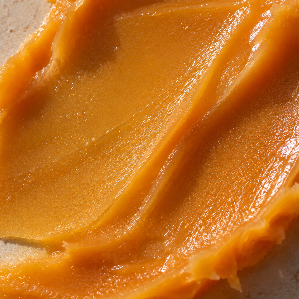 A close-up of a thick, smooth, golden-orange cream or butter spread with glossy highlights on a light beige textured surface.