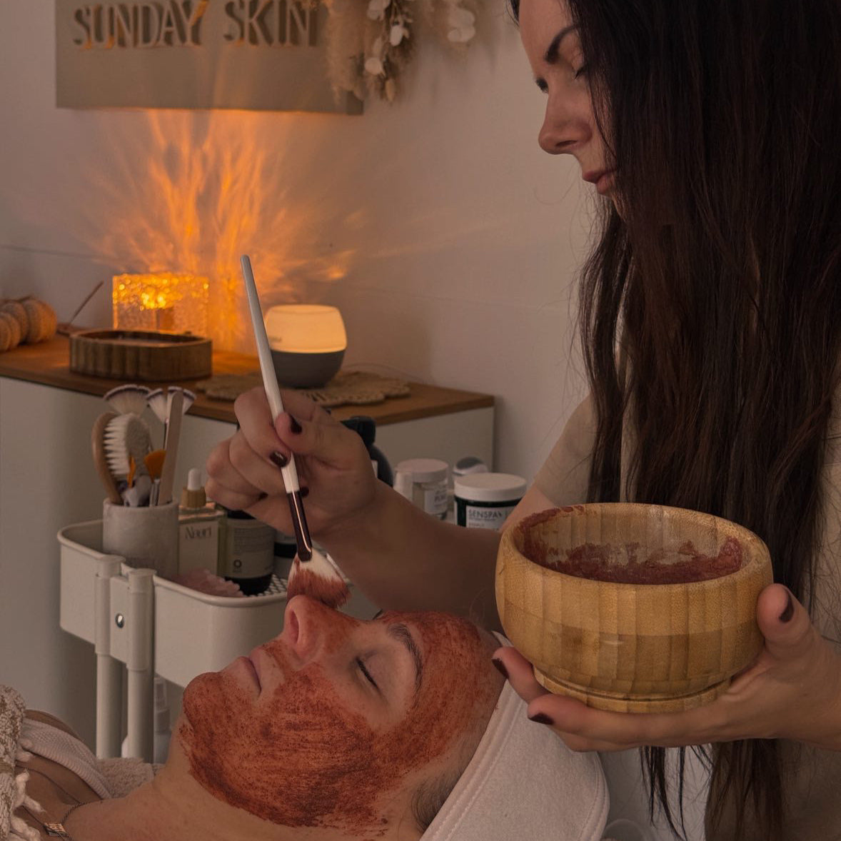 Woman applying a facial mask to a client's face in a spa setting with 'Sunday Skin' branding.