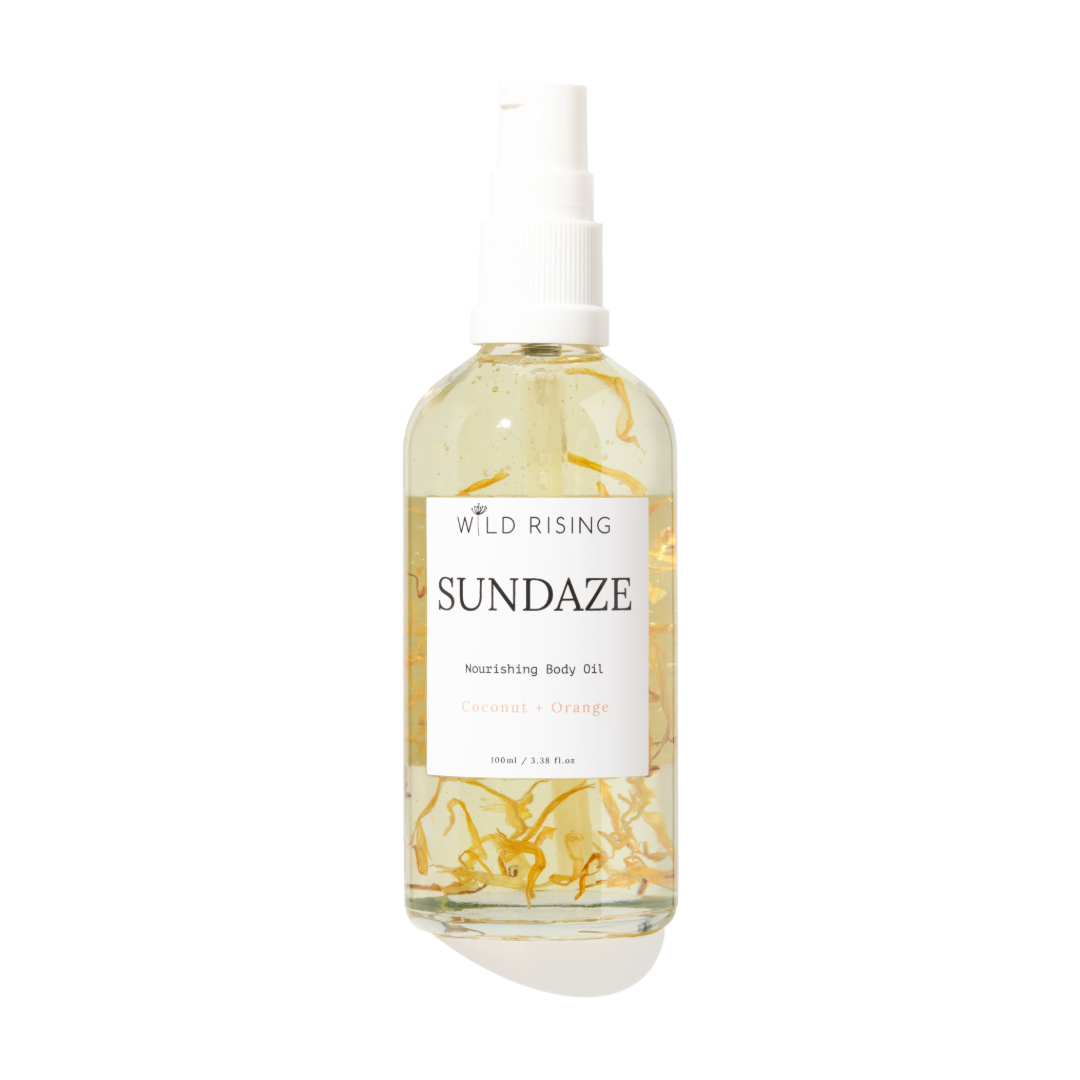 Wild Rising Sundaze Nourishing Body Oil in a clear spray bottle with a white pump dispenser, featuring visible golden-yellow botanical pieces suspended in the oil, set against a plain white background.