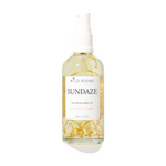 Wild Rising Sundaze Nourishing Body Oil in a clear spray bottle with a white pump dispenser, featuring visible golden-yellow botanical pieces suspended in the oil, set against a plain white background.