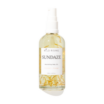 Wild Rising Sundaze Nourishing Body Oil in a clear spray bottle with a white pump dispenser, featuring visible golden-yellow botanical pieces suspended in the oil, set against a plain white background.