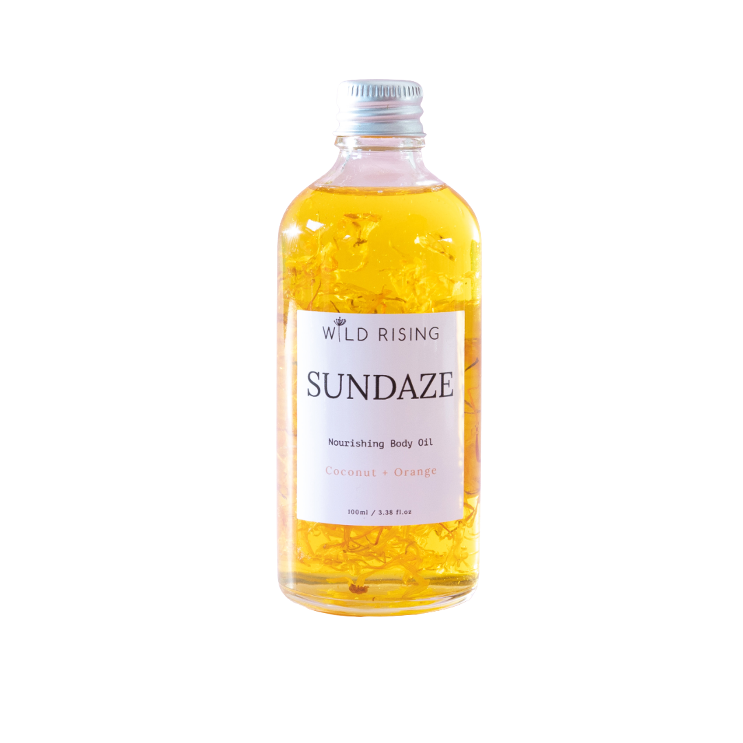 A bottle of Wild Rising Sundaze Coconut + Orange nourishing body oil, filled with golden yellow oil and suspended calendula pieces, on a solid white background.