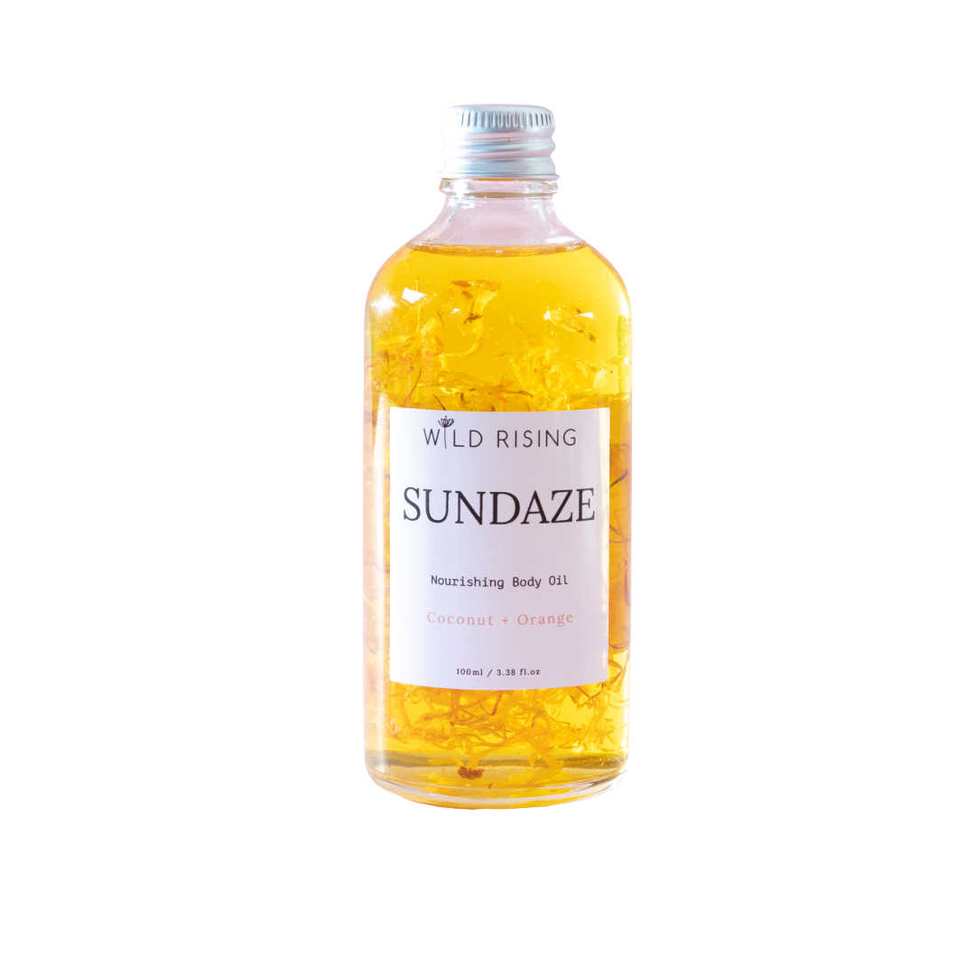 A bottle of Wild Rising Sundaze Coconut + Orange nourishing body oil, filled with golden yellow oil and suspended calendula pieces, on a solid white background.