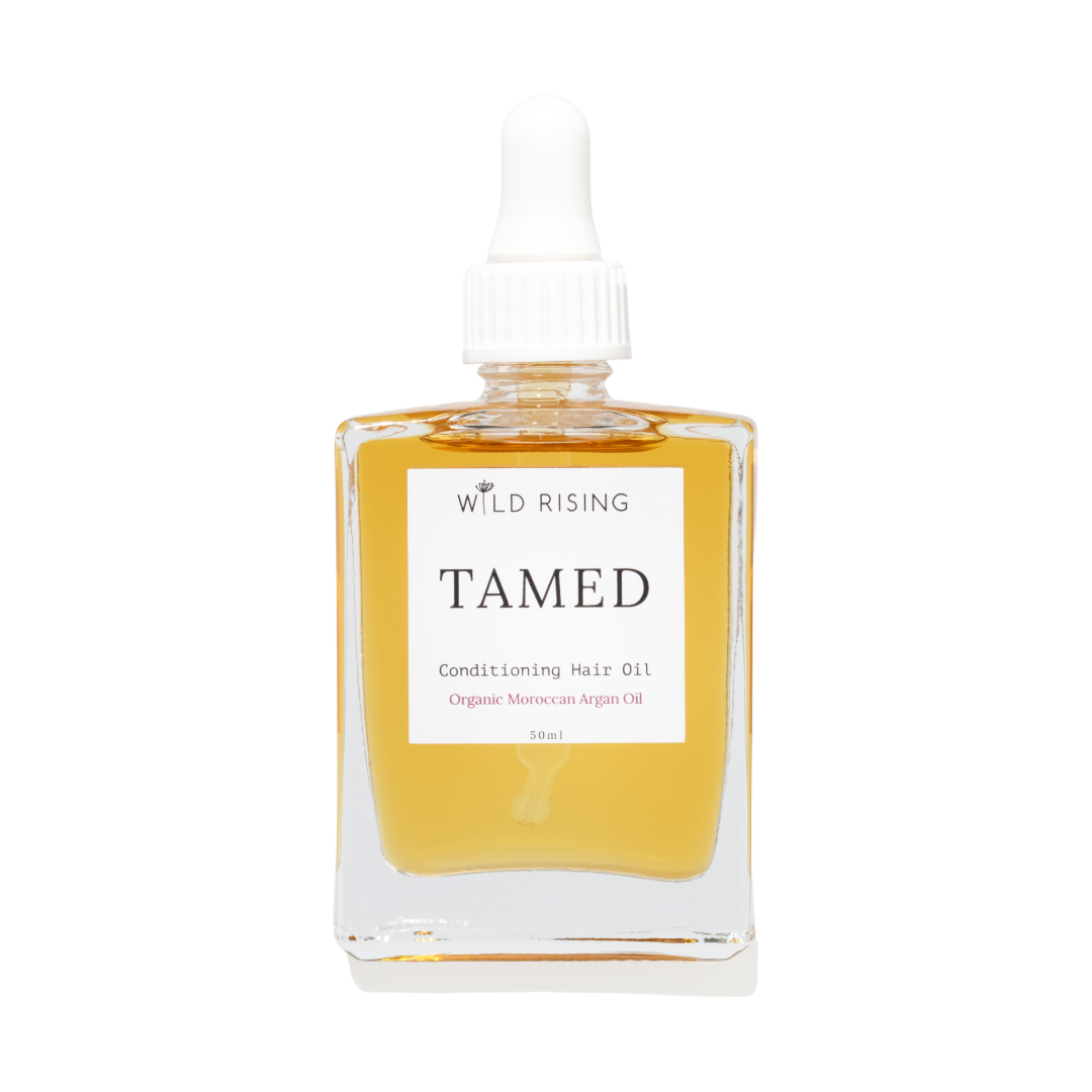 Wild Rising Tamed Conditioning Hair Oil in a clear square glass bottle with a white dropper cap, filled with golden-yellow oil, on a white background.